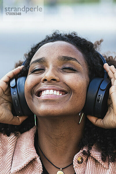 Glückliche Afro-Frau mit geschlossenen Augen hört Musik über Kopfhörer in der Stadt