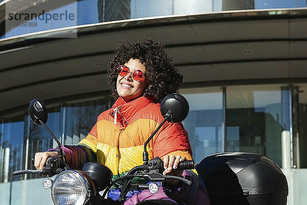 Frau mit Sonnenbrille und bunter Jacke lächelnd auf einem Motorrad sitzend