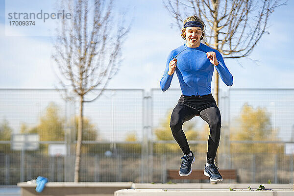 Lächelnder Sportler  der an einem sonnigen Tag auf eine Stützmauer im öffentlichen Park springt