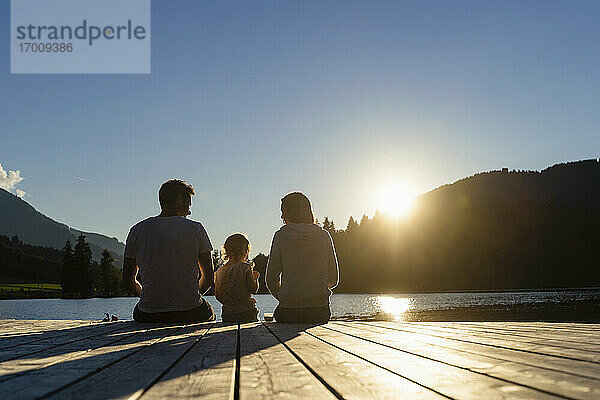 Familie mit kleiner Tochter sitzt bei Sonnenuntergang am Ende eines Stegs am Seeufer zusammen