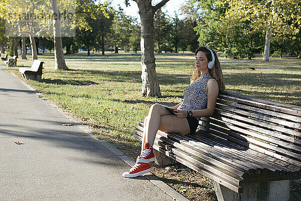 Junge Frau mit geschlossenen Augen hört Musik über Kopfhörer in einem öffentlichen Park an einem sonnigen Tag