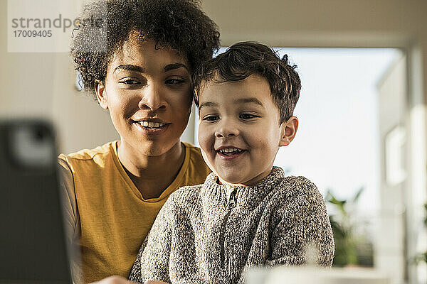 Lächelnde Frau  die ein digitales Tablet benutzt  während sie mit einem Jungen zu Hause sitzt