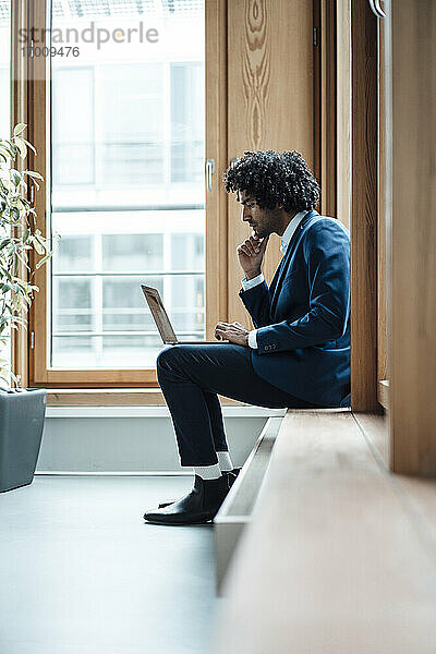 Junger Geschäftsmann benutzt Laptop  während er am Arbeitsplatz am Fenster sitzt