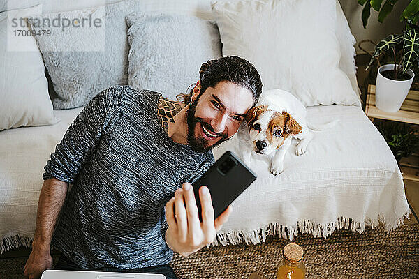 Lächelnder Mann nimmt Selfie mit Hund durch Handy  während er zu Hause sitzt
