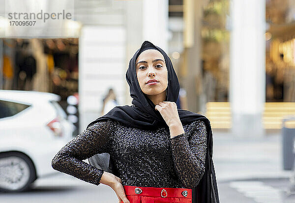 Porträt einer jungen schönen Frau mit schwarzem Hidschab  die mitten auf der Straße mit der Hand auf der Hüfte posiert