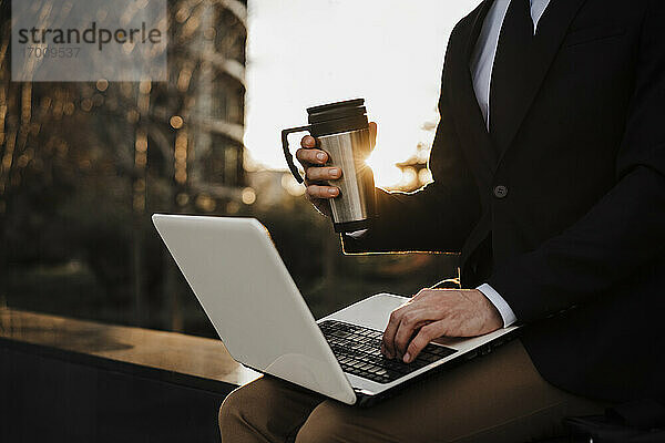 Geschäftsmann mit Thermoskanne  der einen Laptop benutzt  während er bei Sonnenuntergang auf einer Stützmauer sitzt
