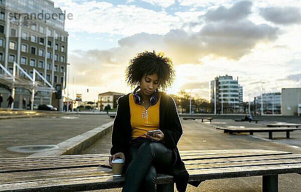 Junge Frau mit Afro-Haar  die ein Mobiltelefon benutzt  während sie auf einer Bank gegen den Himmel in der Stadt sitzt