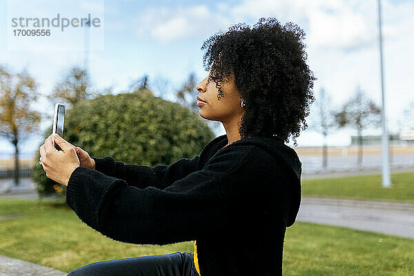 Frau mit Afro-Haar  die ein Selfie über ein Smartphone macht  während sie im Park sitzt