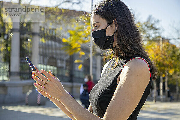 Junge Frau mit Gesichtsmaske  die ein Smartphone benutzt  während sie auf der Straße steht