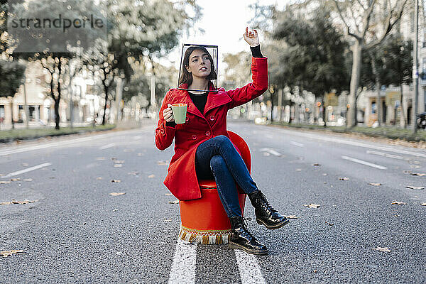 Junge Frau mit Kaffeetasse und Mülleimer auf dem Kopf  während sie auf einem Stuhl auf der Straße sitzt