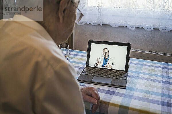 Ein älterer Mann lässt sich von einem männlichen Allgemeinmediziner per Video über einen Laptop im Wohnzimmer beraten