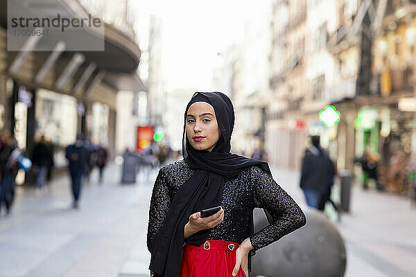 Porträt einer jungen Frau mit schwarzem Hidschab  die mit einem Smartphone in der Hand auf dem Gehweg steht