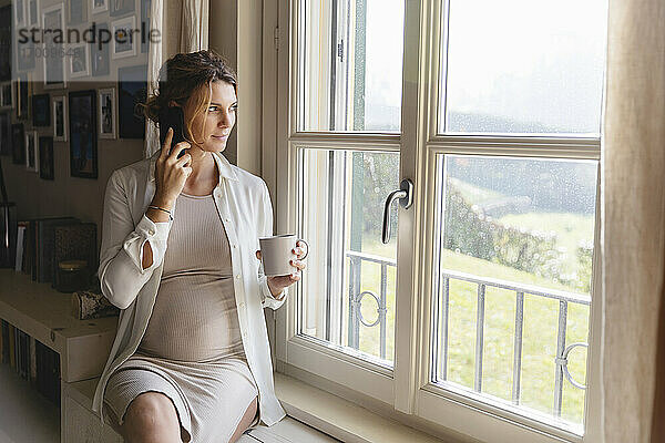 Schwangere Frau  die mit ihrem Handy telefoniert und durch ein Fenster schaut