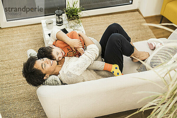 Lächelnde Mutter  die ihren Sohn umarmt  während sie sich zu Hause auf dem Sofa ausruht