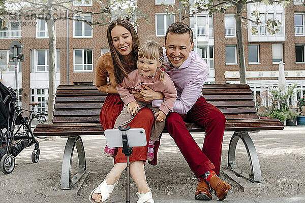 Glückliche Familie mit einer Tochter  die ein Selfie mit ihrem Handy auf einem Stativ auf einer Parkbank macht