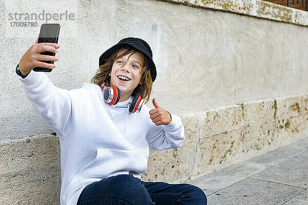 Junge mit Hut und Kopfhörern  der ein Mobiltelefon benutzt  während er an der Wand sitzt