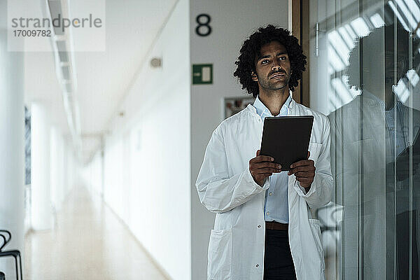 Nachdenklicher junger Arzt  der ein digitales Tablet hält und sich an eine Glaswand im Krankenhausflur lehnt