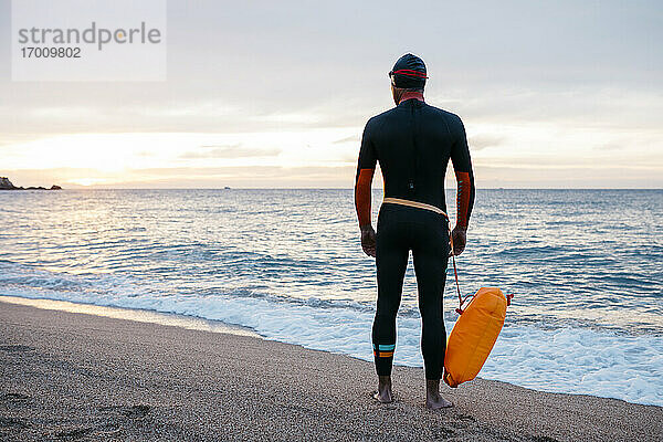Männlicher Schwimmer  der allein am sandigen Küstenstrand steht  mit Schwimmflügel in der Hand