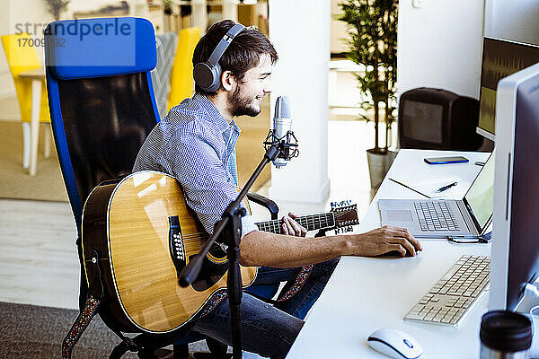 Männlicher Musiker mit Kopfhörern und Gitarre  der lächelnd einen Laptop im Aufnahmestudio benutzt