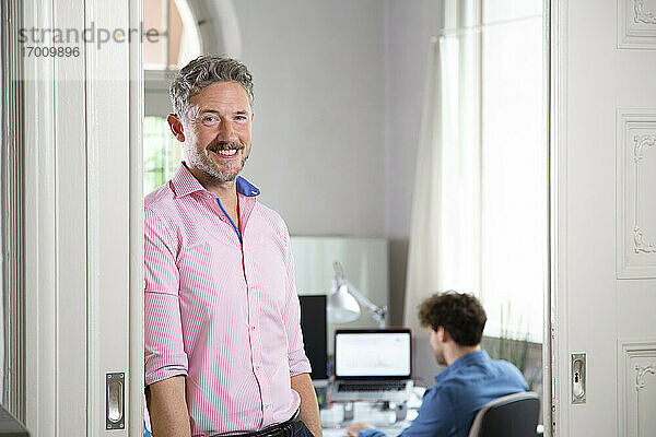 Lächelnder Geschäftsmann mit einem männlichen Kollegen im Hintergrund in einer Bürokabine
