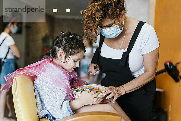 Friseurin und Mädchen suchen im Korb mit Haarspangen beim Friseur während des Coronavirus