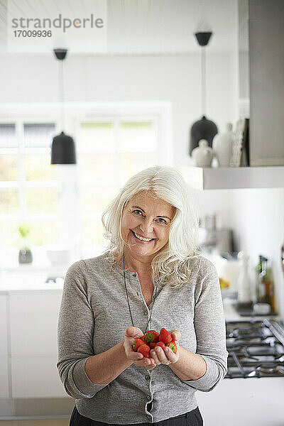 Lächelnde Frau im Ruhestand mit frischen Erdbeeren in der Hand  während sie in der Küche zu Hause steht