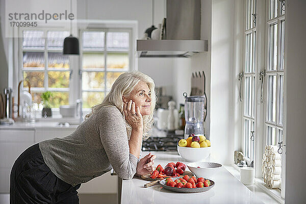 Nachdenkliche Rentnerin lehnt sich mit Erdbeeren in der Wohnung an den Küchentisch