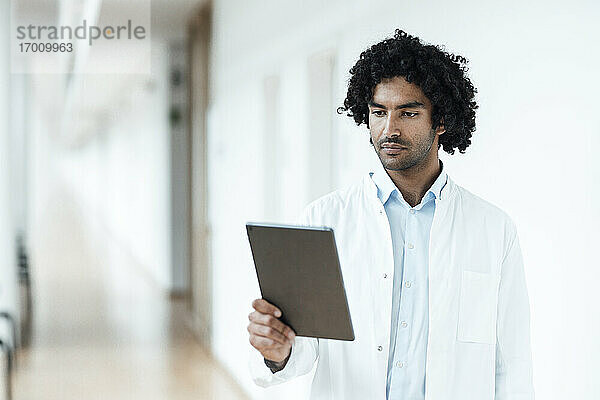 Selbstbewusster junger Arzt  der ein digitales Tablet auf dem Flur eines Krankenhauses benutzt