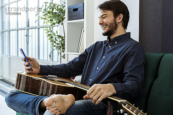 Lächelnder Musiker  der ein Mobiltelefon benutzt  während er mit seiner Gitarre im Studio sitzt