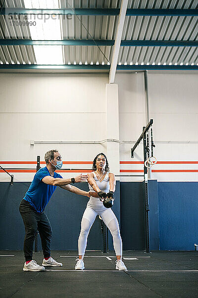 Männlicher Fitnesstrainer trainiert eine Sportlerin mit einer Kettlebell im Fitnessstudio während COVID-19