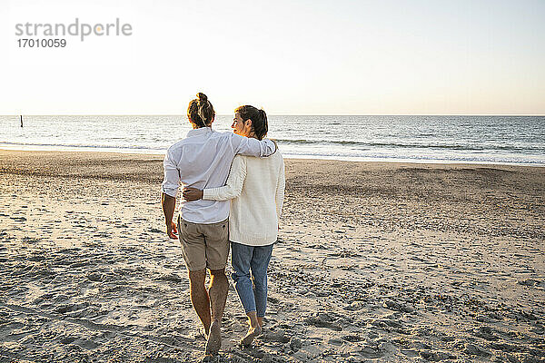 Junges Paar  das sich in den Armen liegt und bei Sonnenuntergang am Strand spazieren geht