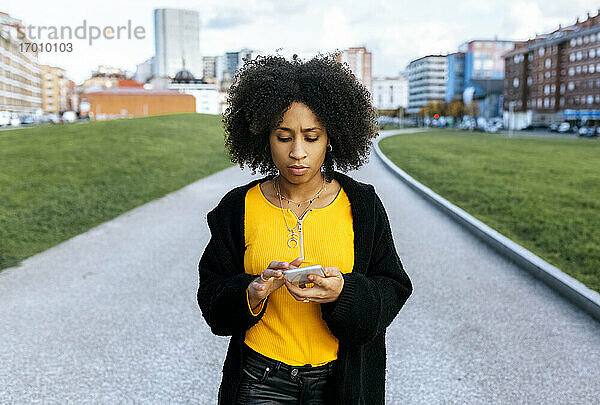 Junge Frau mit Afro-Haar  die ein Mobiltelefon benutzt  während sie auf einem Fußweg in der Stadt steht
