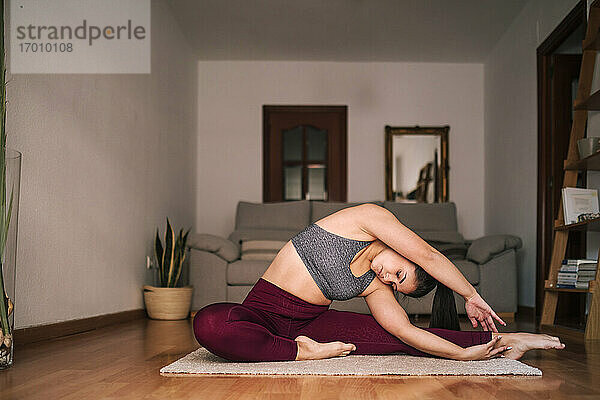 Junge Frau mit erhobenen Armen übt Yoga  während sie zu Hause auf einer Matte sitzt