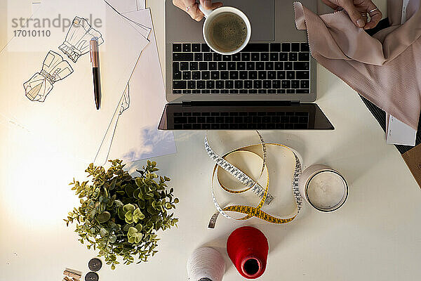 Modedesigner hält Kaffeetasse über Laptop mit Stoffmuster am Schreibtisch im Atelier