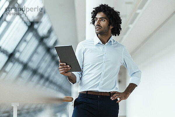 Nachdenklicher männlicher Unternehmer  der ein digitales Tablet in der Hand hält  während er mit der Hand auf der Hüfte am Geländer im Flur eines Büros steht
