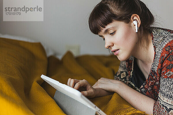 Junge Frau  die auf dem Bett liegend einen Online-Kurs über ein digitales Tablet mit drahtlosen Kopfhörern verfolgt