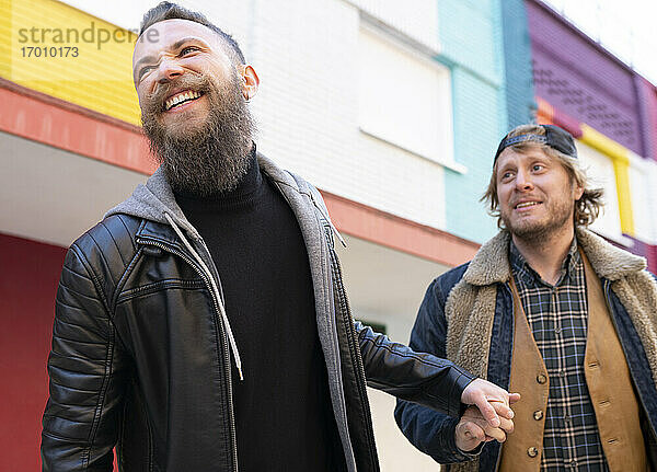 Lächelnde Homosexuell Paar hält die Hände beim Gehen gegen bunte Gebäude in der Stadt