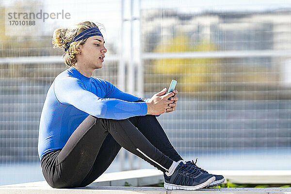 Blonder männlicher Sportler  der in einem öffentlichen Park an einem sonnigen Tag ein Mobiltelefon benutzt