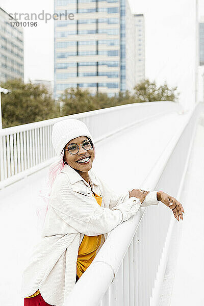 Lächelnde Frau mit Perücke und Mütze auf einer Brücke stehend