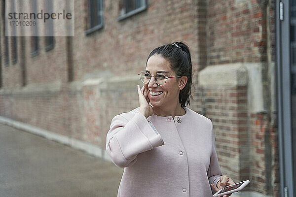 Lächelnde Geschäftsfrau  die ihr Smartphone in der Hand hält und gegen ein Gebäude blickt