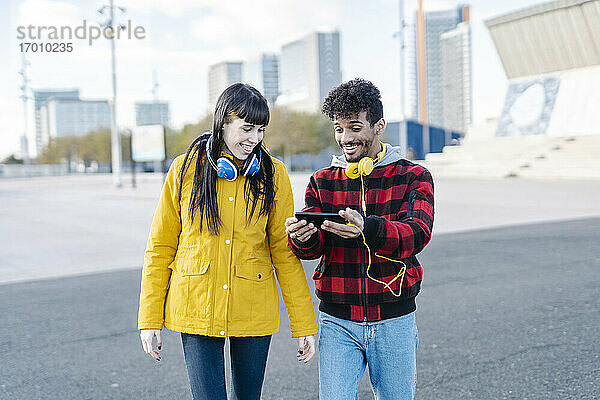 Lächelndes Paar mit Kopfhörern und Mobiltelefon beim Gehen auf der Straße