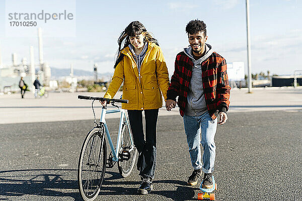 Lächelnder Mann fährt Skateboard und hält die Hand eines Freundes  der mit dem Fahrrad auf der Straße fährt