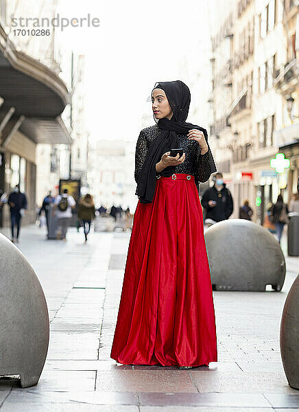 Porträt einer jungen Frau mit schwarzem Hidschab  die mit einem Smartphone in der Hand auf dem Gehweg steht