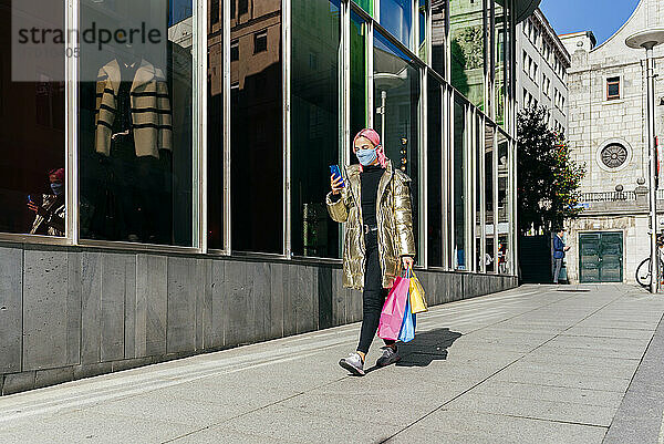 Frau mit Gesichtsschutz  die ein Mobiltelefon benutzt  während sie mit Einkaufstaschen auf dem Gehweg geht