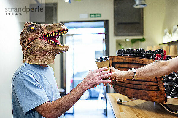 Hand eines Barista  der einem Mann mit Dinosauriermaske in einem Café Kaffee serviert