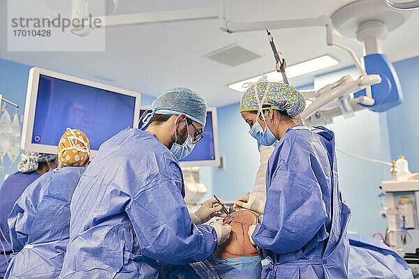 Ärzte mit Gesichtsmaske  die eine Operation durchführen  während sie mit einem Kollegen im Hintergrund im Operationssaal stehen