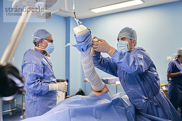 Arzt wickelt Hand  während er mit einem Kollegen im Hintergrund im Operationssaal steht