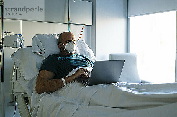 Männlicher Patient mit Gesichtsmaske bei der Arbeit am Laptop  während er im Krankenhaus auf dem Bett sitzt