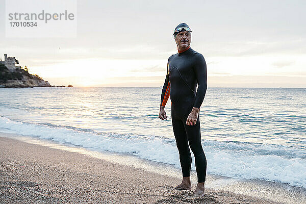 Porträt eines männlichen Schwimmers  der bei Sonnenuntergang allein am Küstenstrand steht