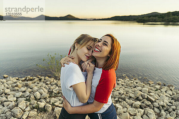 Glückliche Frau  die ihre Freundin umarmt  während sie bei Sonnenuntergang am See steht